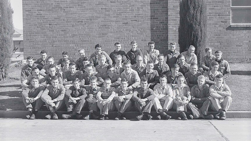 A black and white photograph of men in overalls and 1950s clothes outside a brick building