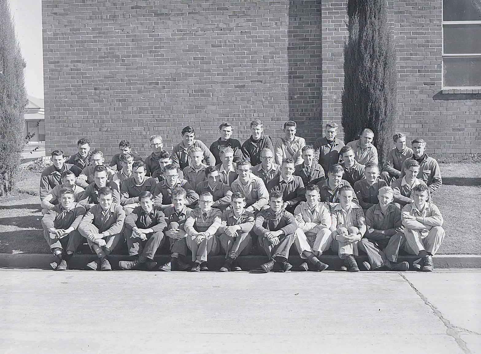 A black and white photograph of men in overalls and 1950s clothes outside a brick building