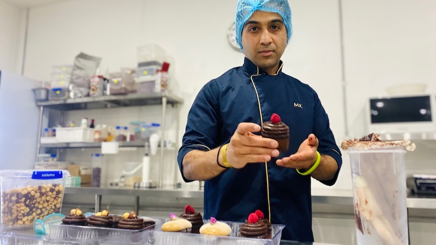 A business owner in a hair net in a commercial kitchen holds a cup cake and looks at the camera 