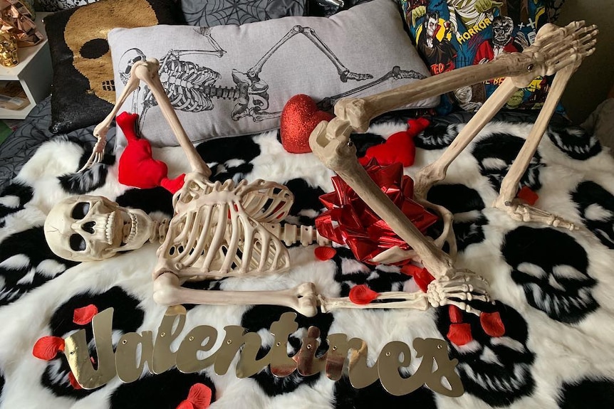 A skeleton suggestively posing on a bed, surrounded by rose petals.