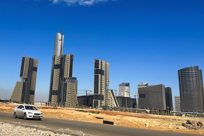 A car drives down a road next to a desert landscape with skyscrapers in the background against a bright blue sky