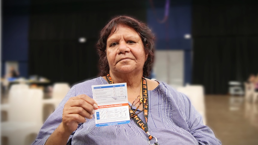 Indigenous woman in blue stripy shirt sits in chair in big hall holding up vaccination certificate