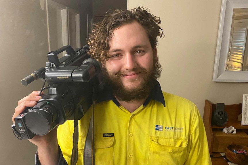 An image of a man with short brown curly hair holding an old video camera, the technology is out-of-date.