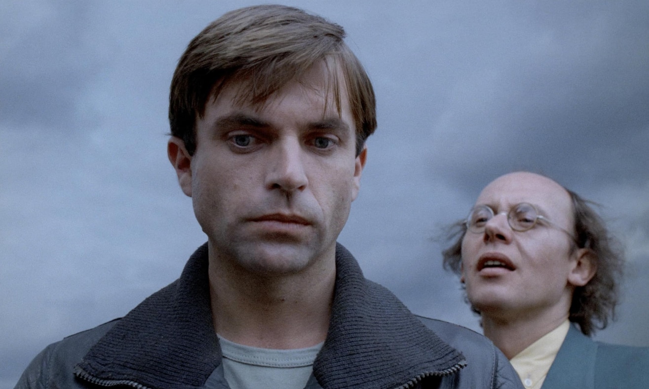 Film still of a young Sam Neill looking pensive, behind him an odd looking man and a gloomy sky