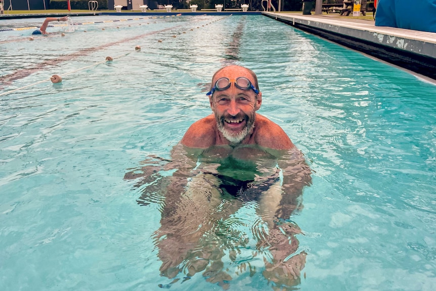 A man, with goggles on his head, smiles in a public swimming pool.