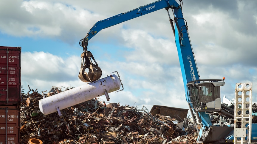 A blue crane lifts a large white container onto a pile of scrap metal.