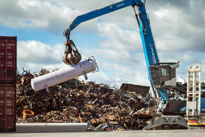 A blue crane lifts a large white container onto a pile of scrap metal.