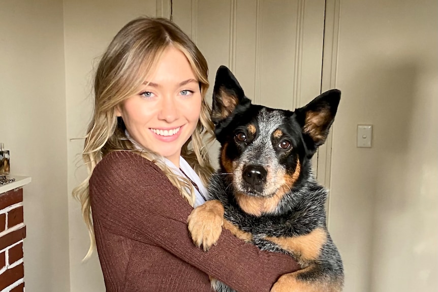 A smiling young blonde woman cuddles her kelpie.