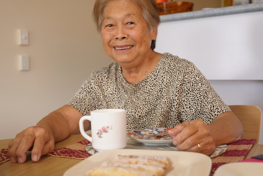 Gaby Nagel sits at a dining table smiling, cup of tea, fresh spring rolls in the foreground. 