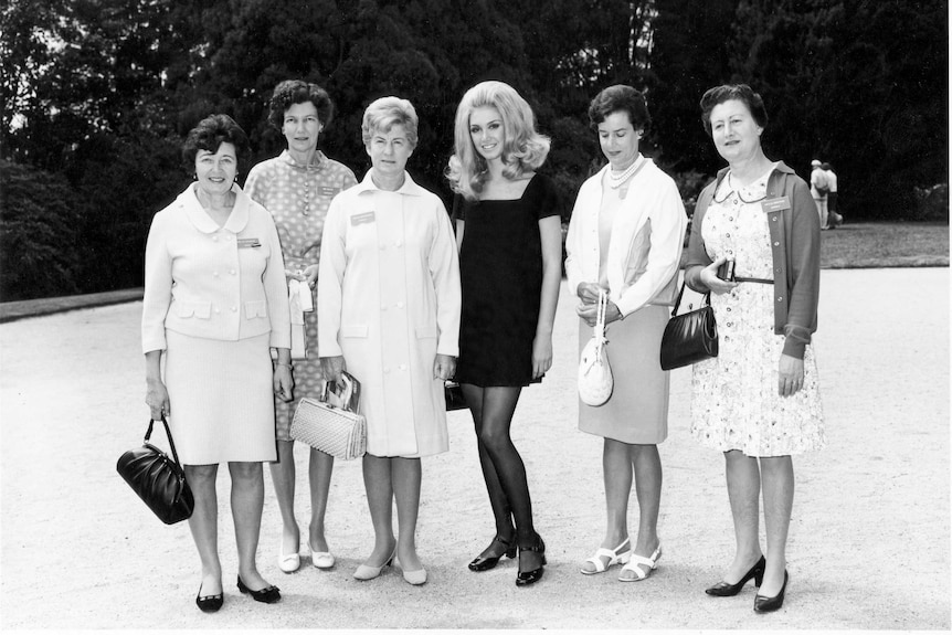 A woman in a black minidress stands among women in conservative outfits, one looking at the ground.
