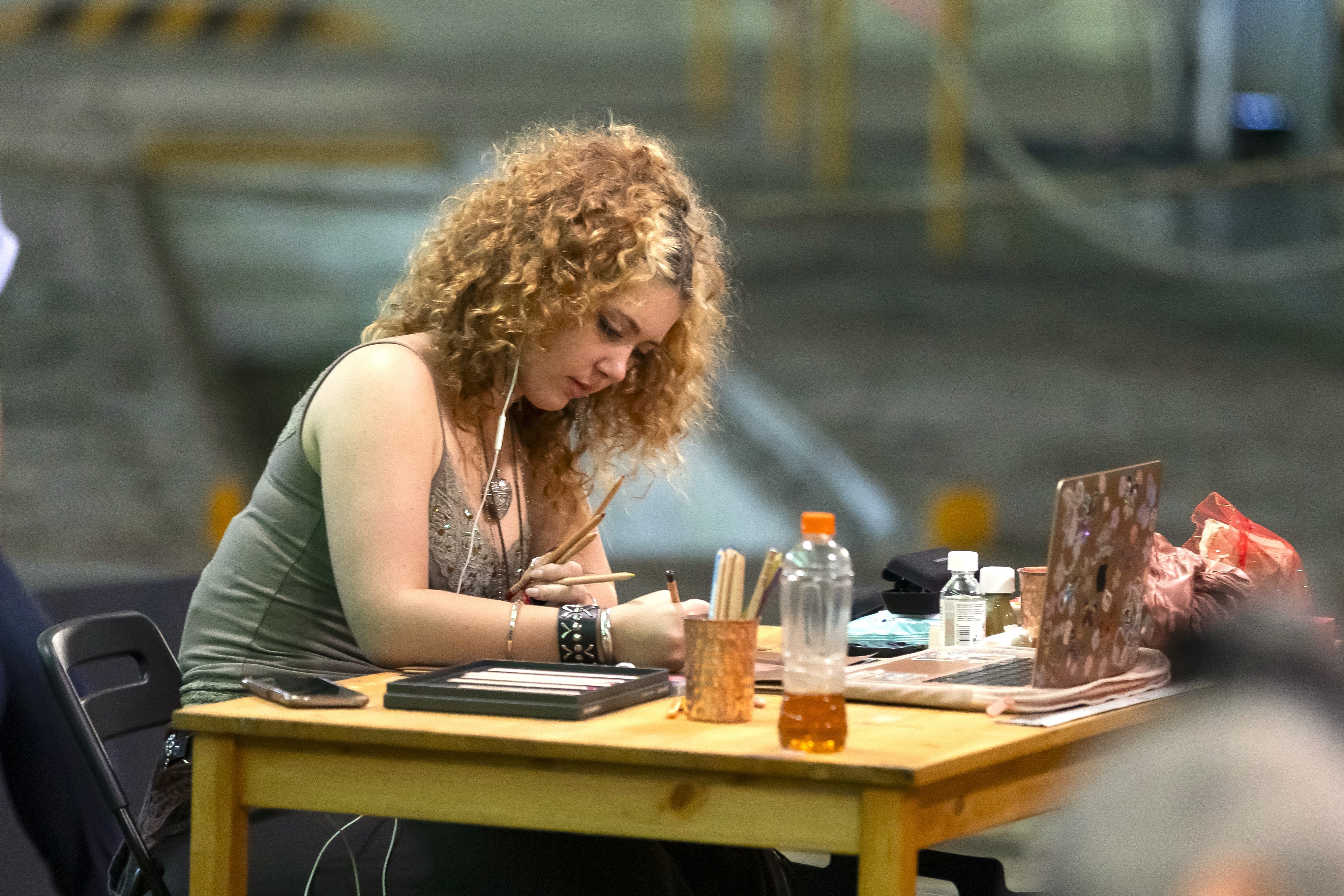 A young woman with curly hair stares down at a crowded table with multiple pencils in her hand as she draws, one headphone in