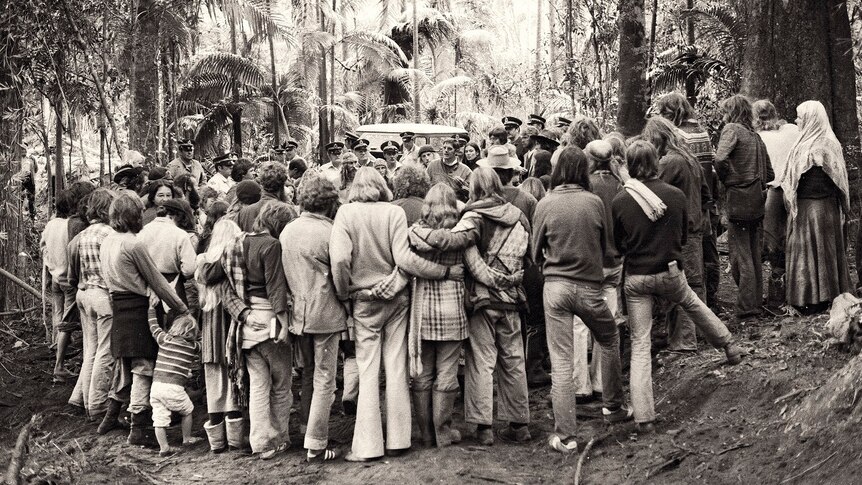 A black and white photo of a large group of people standing in a rainforest talking to police.