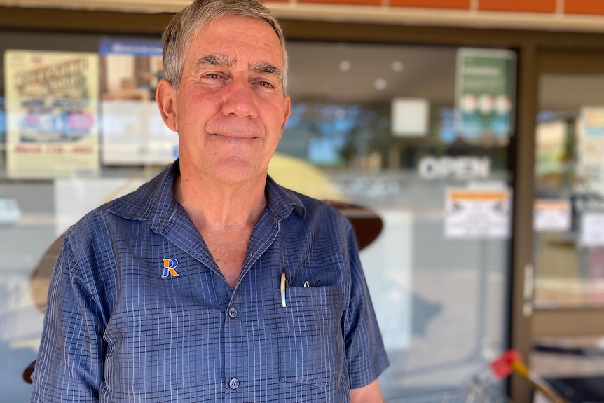 A man with greying hair has a slight smile, he wears a blue shirt with an op shop sign in the background