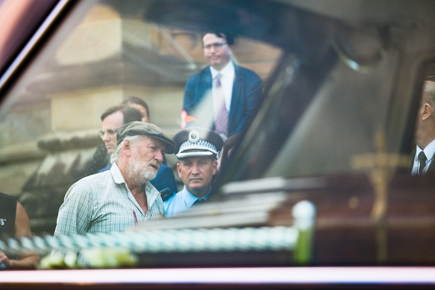 A man in a hat speaks to a police officer in uniform, seen through the window of a hearse carrying a casket