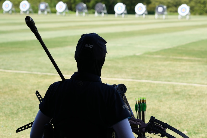 An archer is pictured in the shadows at one end of a range, with a row of archery targets standing in the distance.