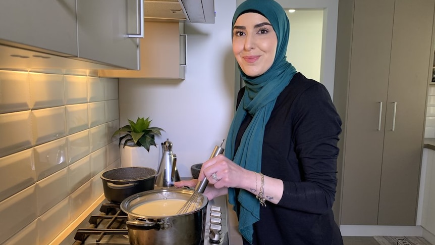 A woman stands in her kitchen and smiles at the camera stirring food in a pot.