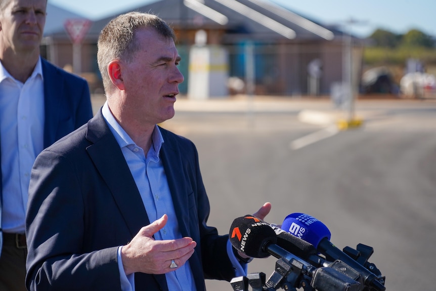 A man in a suit and blue shirt addresses the media, a house being built is in the background