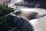 An aerial view of a dam wall with a crane on it and workers' trucks in the background.