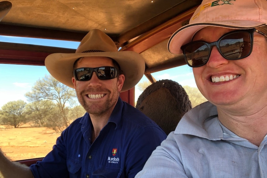 Man and woman in vehicle smile at the camera, as they drive through hot dry pastoral country in WA. 