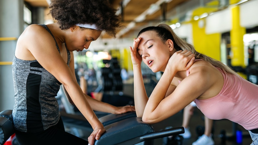 Two women leaning on an exercise machine looking exhausted. One is wearing a black and grey singlet. The other has a pink sing