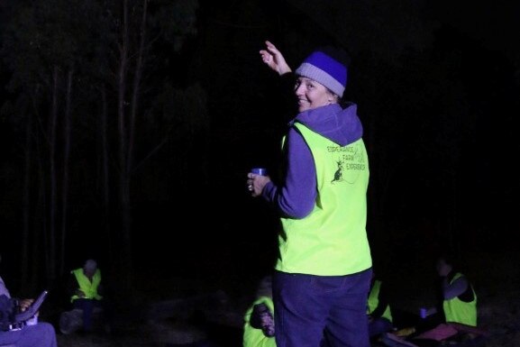 Woman in high-vis holds up her hand, gesturing to people and smiling. The sky is dark.