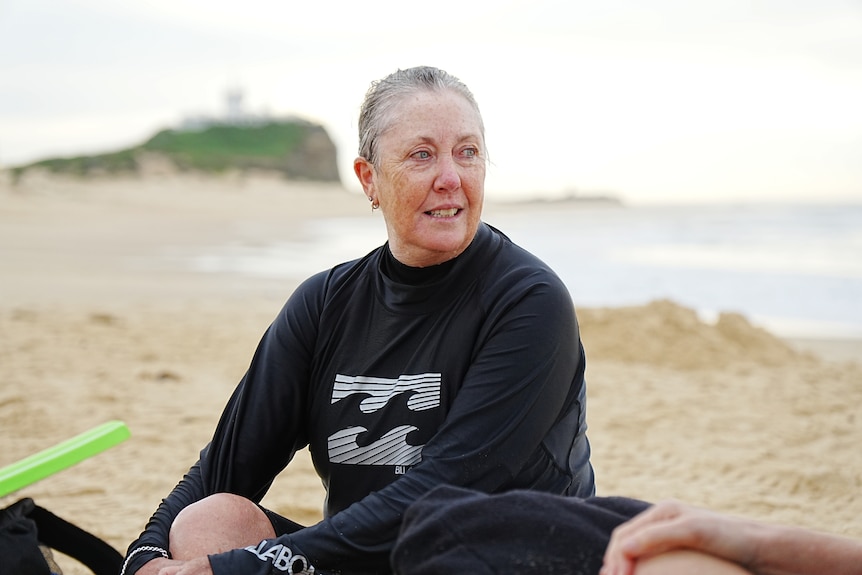 A woman with grey hair, wearing a wetsuit and sitting on sandy beach.