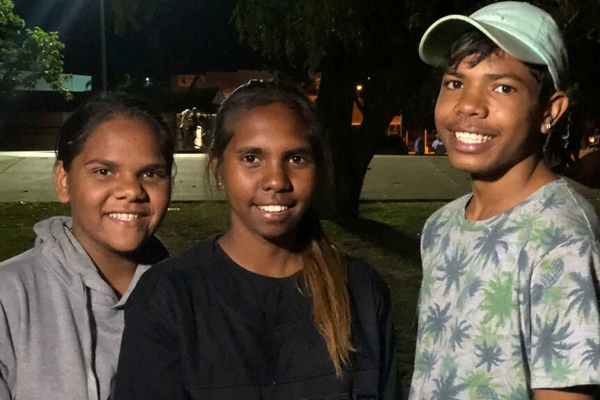 Two Aboriginal teenage girls and one Aboriginal teenage boy at an outdoor location at night time, smiling at camera