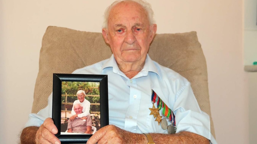 Len sitting on a sofa, wearing his medals, holding a framed photo of him and his wife.