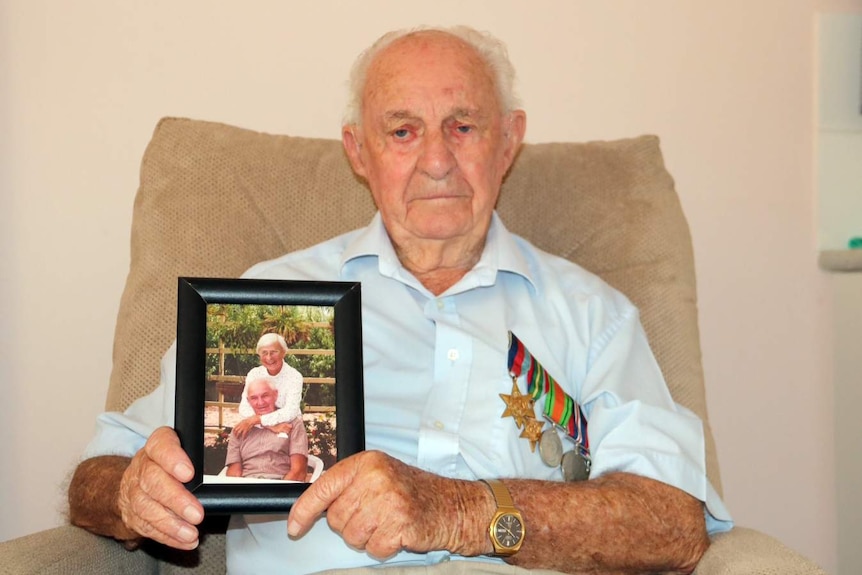 Len sitting on a sofa, wearing his medals, holding a framed photo of him and his wife.