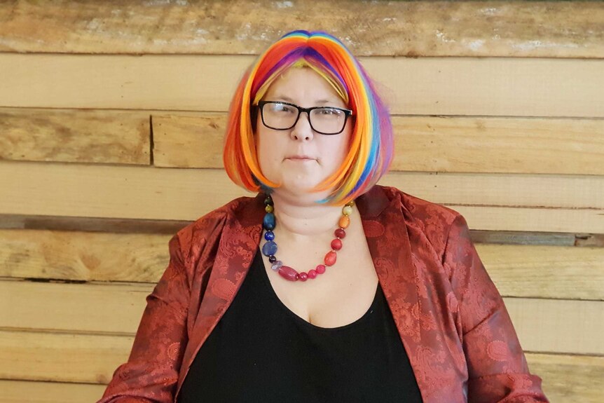 A person with long multi-coloured hair sitting at a table wearing a brown jacket and glasses.