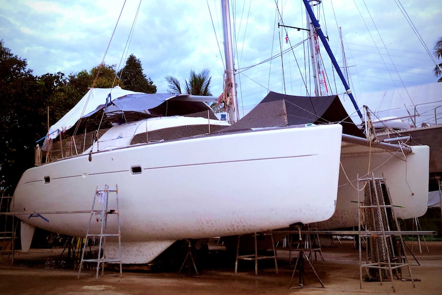 A white boat with mast up on concrete at a boatyard. Photo taken from starboard bow.