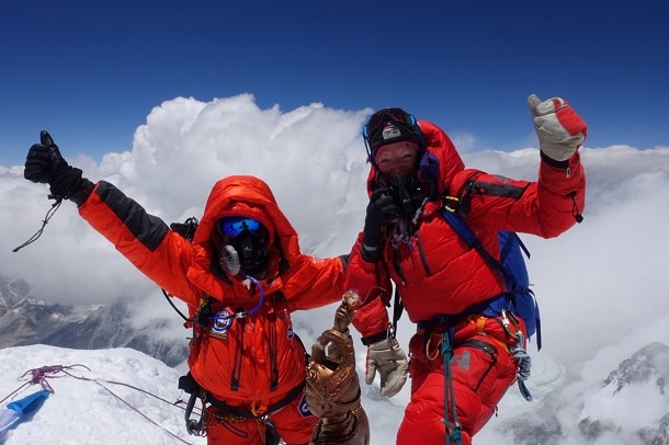 Two female mountain climbers in red outfits raise their arms on the summit of Mount Everest