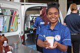 A teenage girls holds up a cup of coffee in a paper cup in front of a mobile coffee van