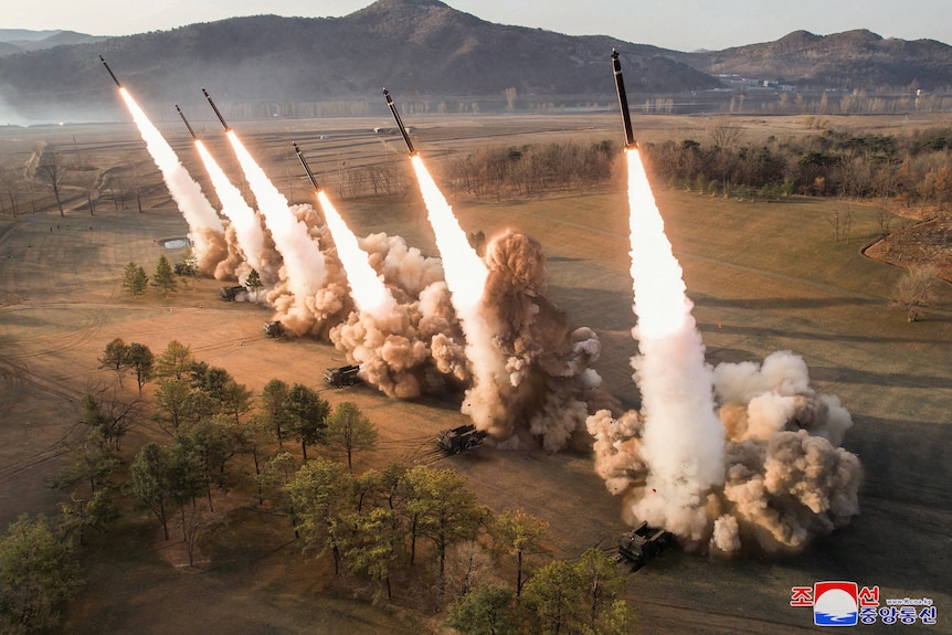 Six large missile blast off from launch systems arranged over brown open land with a mountain behind.