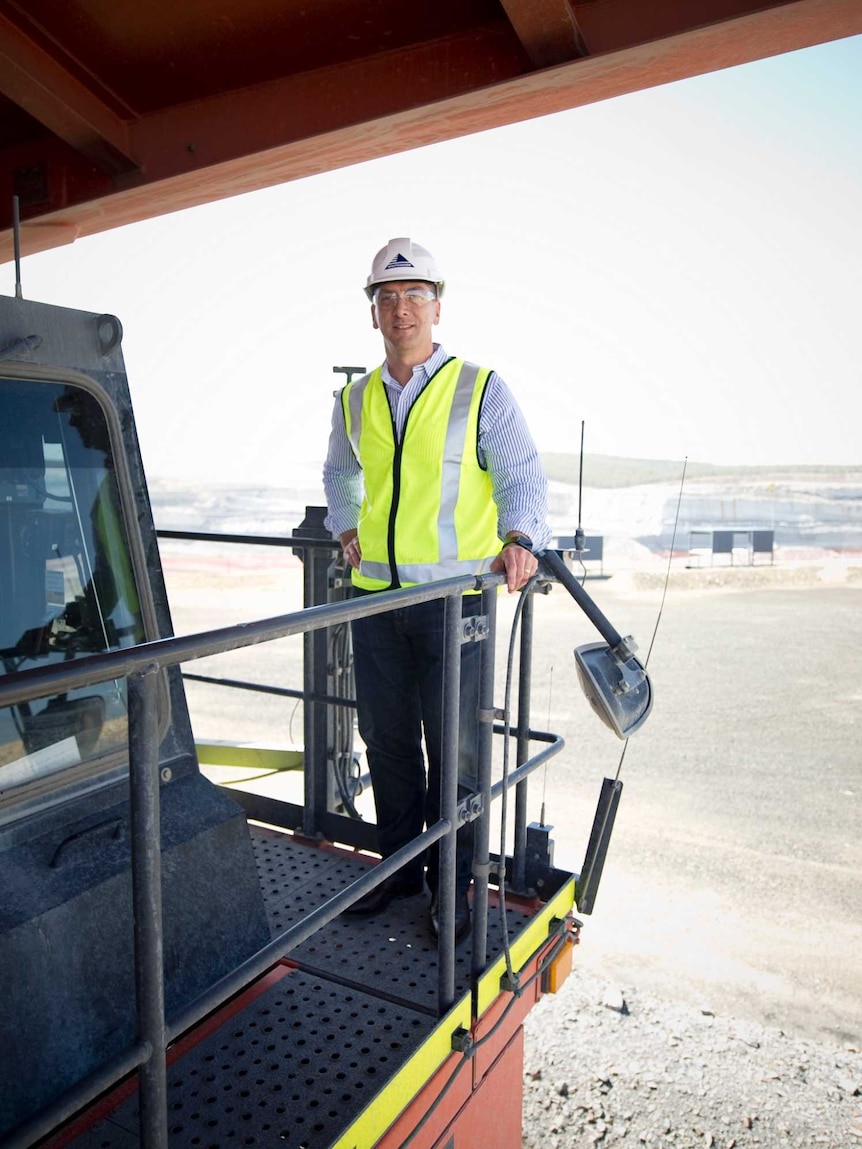 A man standing on a truck wearing a high-vis vest and hard hat with a coalfield in the background on a clear day