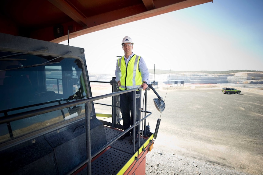 A man standing on a truck wearing a high-vis vest and hard hat with a coalfield in the background on a clear day.