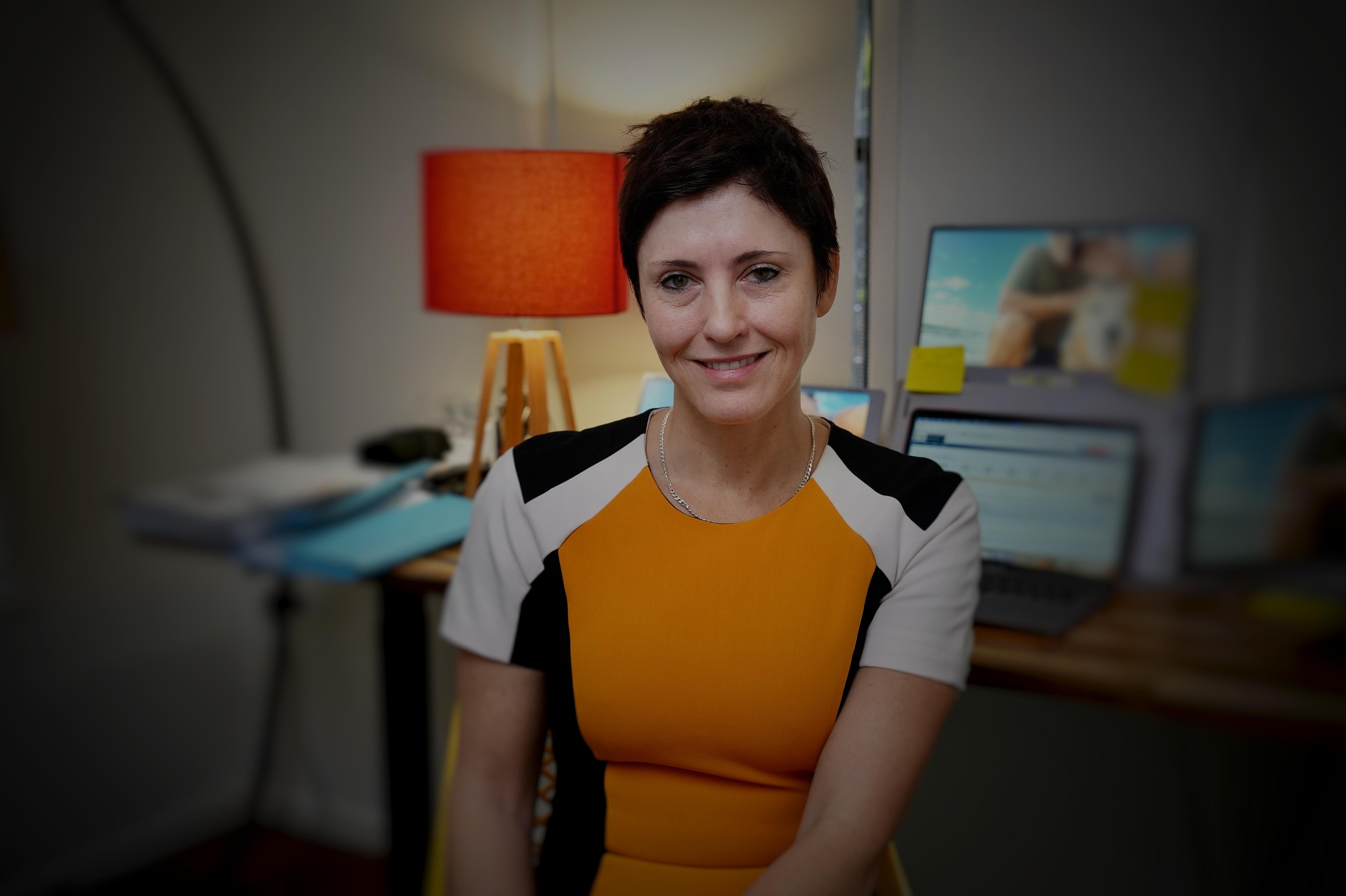 A white woman with a black pixie cut sitting at her office desk wearing orange, white, black top looking right of frame.