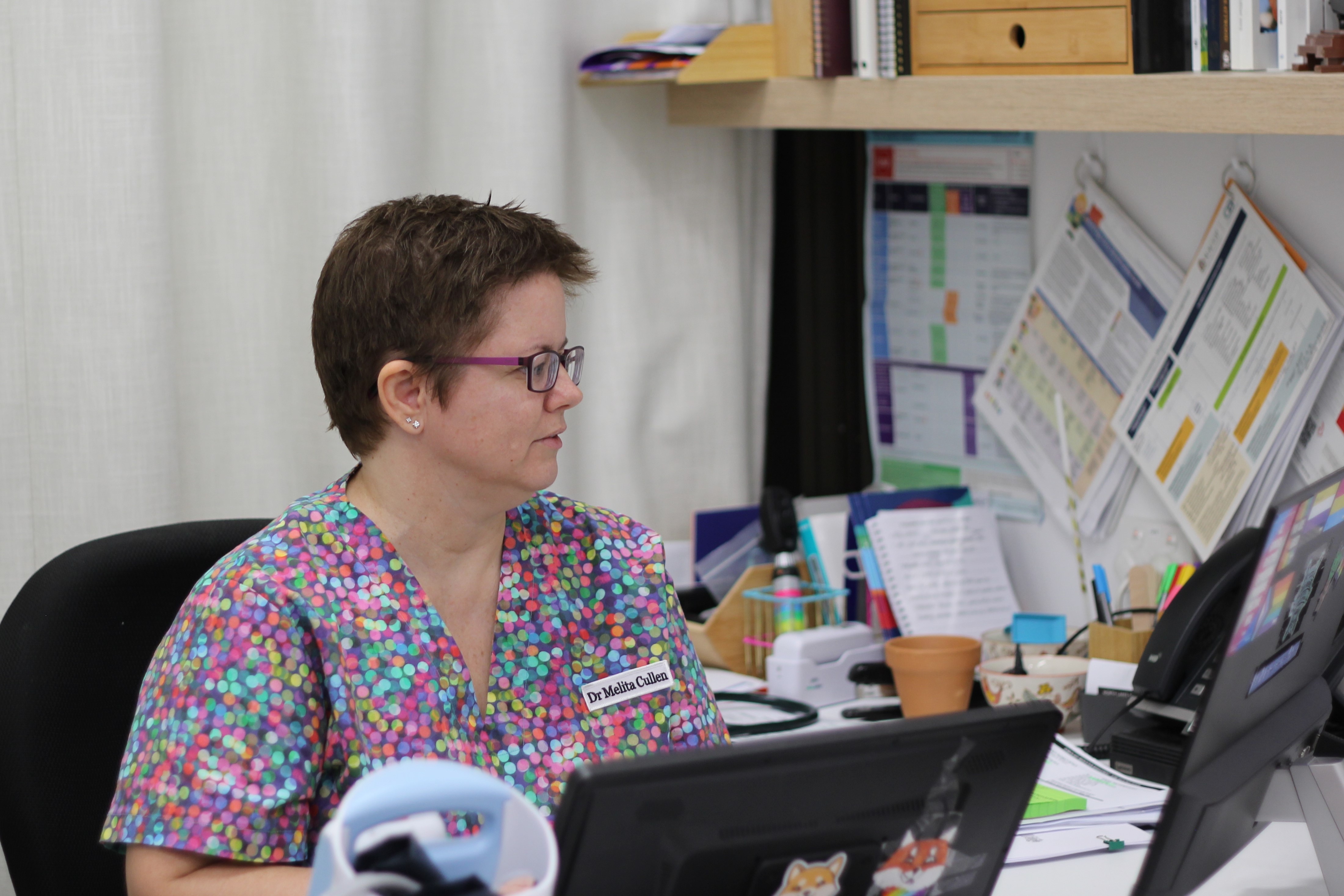 A woman with short brown hair and glasses and colourful scrubs sits at a computer desk