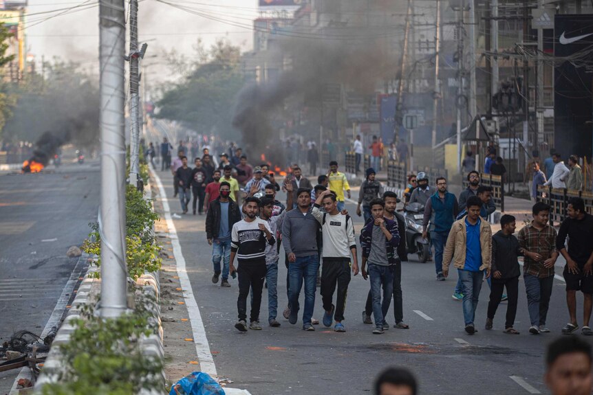 A crowd of protestors walk past burning tires in a main street as smoke rises behind them.