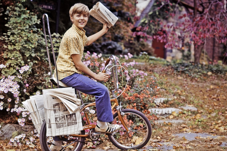 一名报童穿着球鞋，骑着自行车送报纸。