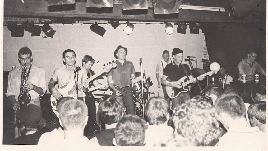 A black and white scanned photo of Strange Tenants on stage. We see a saxophonist, guitarists, vocalist and drummer.