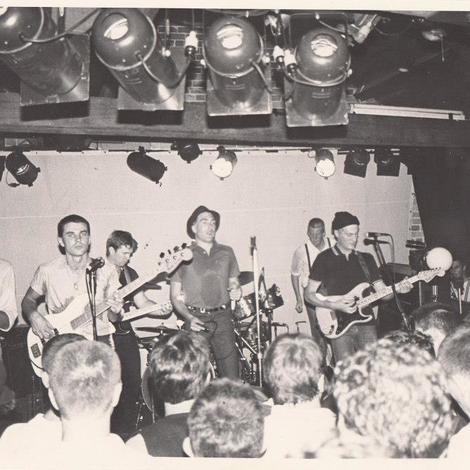 A black and white scanned photo of Strange Tenants on stage. We see a saxophonist, guitarists, vocalist and drummer.