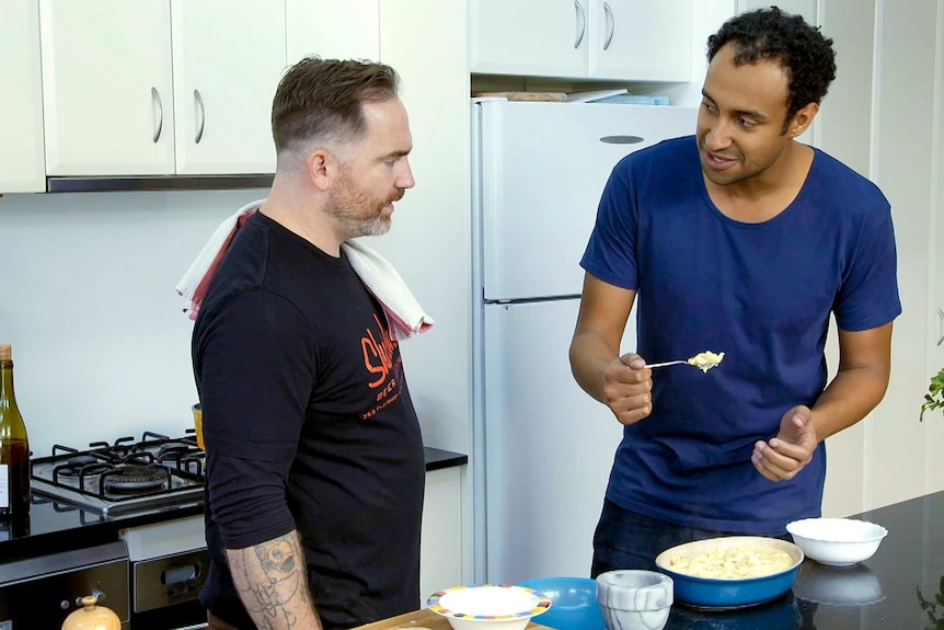 Two men in the kitchen next to a dish full of mac and cheese.