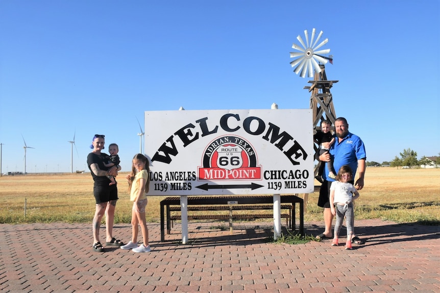 A family of six stand beside a road sign in the United States. A mum, a dad who are each holding babies, two girls