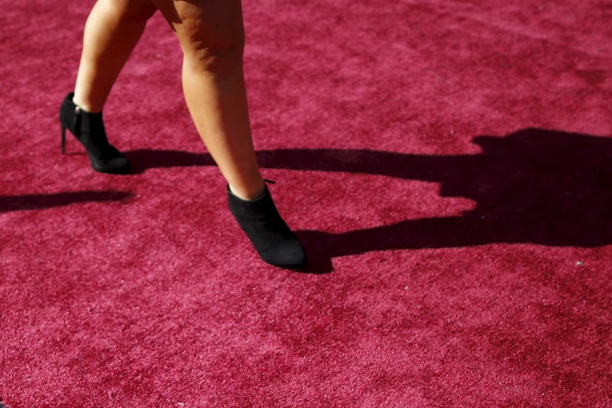 A woman in black high heel shoes steps on the Oscars carpet, which is the trademarked shade of red.