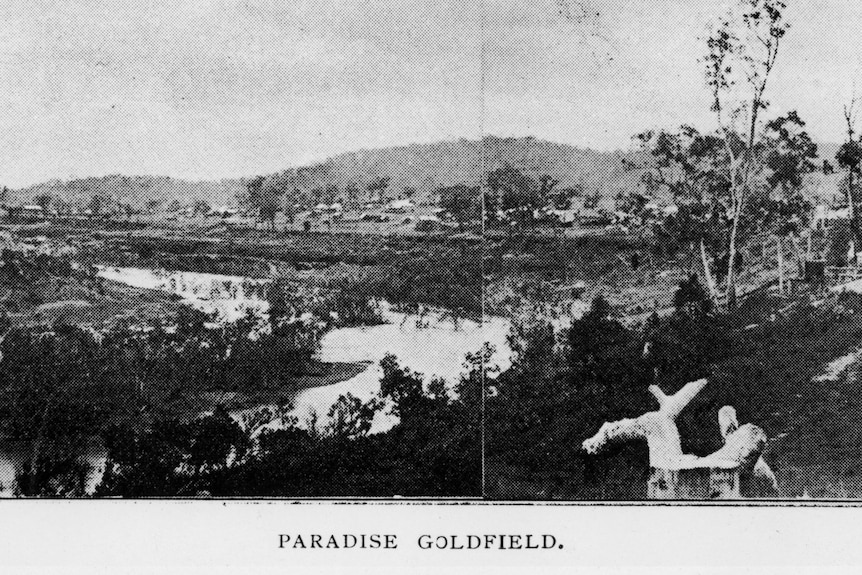 A grainy black and white image of distant small buildings across a bend in a river.