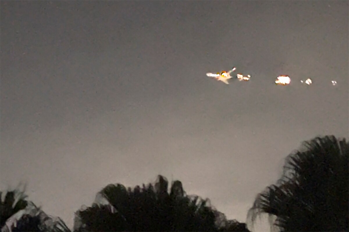 A screenshot from a video showing tops of trees in the foreground and a plane on fire in the sky in the background, at night