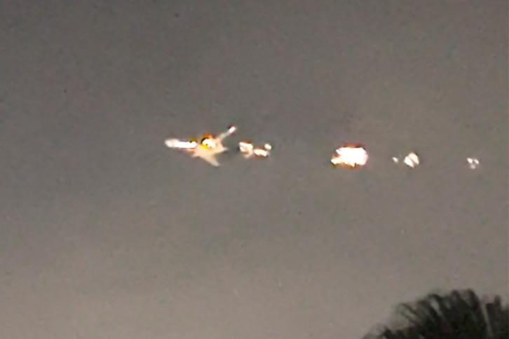 A screenshot from a video showing tops of trees in the foreground and a plane on fire in the sky in the background, at night