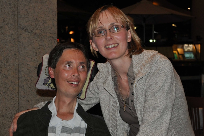 Michael Lee, sitting in his wheelchair, looks at his wife, Joanna, who stands next to him with her arm around Michael.