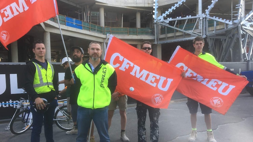 Union members stand with red and white CFMEU flags in front of a building site.